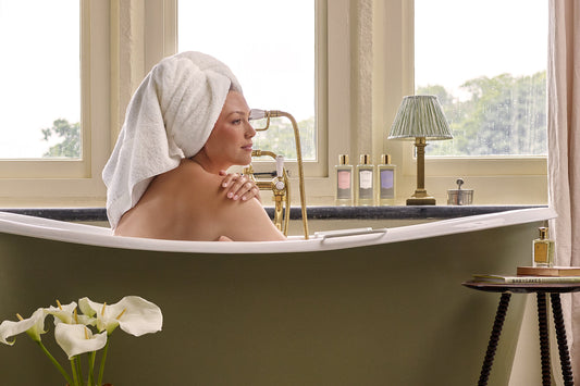 A woman sat in a green bath, in front of a large brightly lit window. She has Floris Bath & Shower Gel on the shelf and a bottle of Stephanotis Bath Essence purched on a wooden stool.