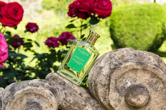 A bottle of Floris London's Rose Geranium fragrance, balanced on a stone wall in a sunlit garden with rose bushes.