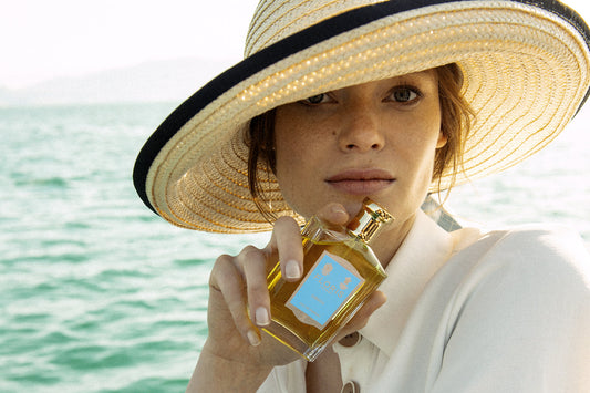 A lady holding a bottle of Sirena, she is wearing a large hat and is sat infront of the waters edge.