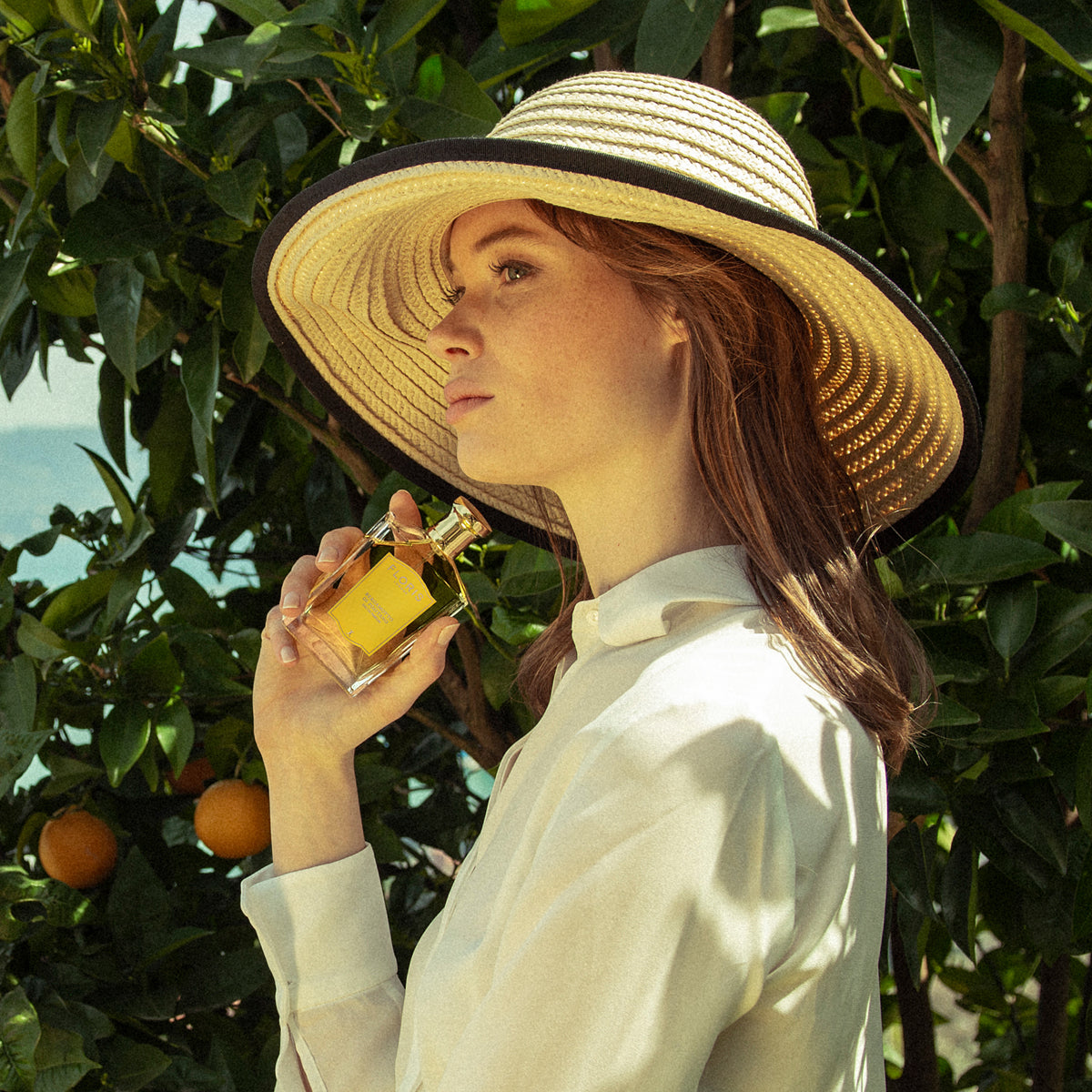 A woman in a straw hat holds Floris London's Bergamotto di Positano, with an orange tree and blossoms in a lush garden.