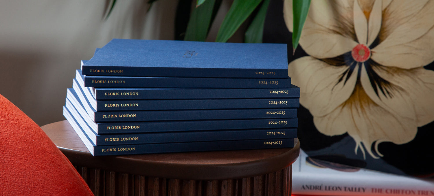 A pile of navy Floris London 2024/25 catalogue upon a wooden table and next to a vase with a painting of a flower.