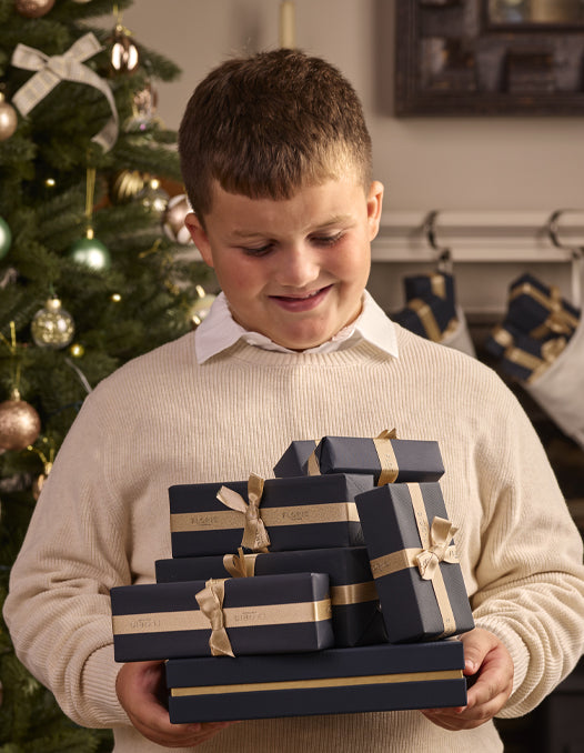 A boy in a cream jumper, standing in front of a decorated Chirstmas tree holding a pile of Floris London luxury gift boxes.