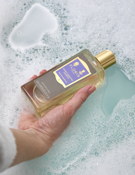 A hand holding a bottle of Night Scented Jasmine Bath & Shower Gel just above the surface of a bubble-filled bath.