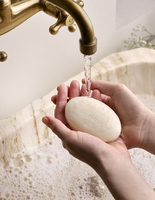 Hands holding a luxurious Floris London soap under a running brass tap, with a sink filled with bubbles beneath.