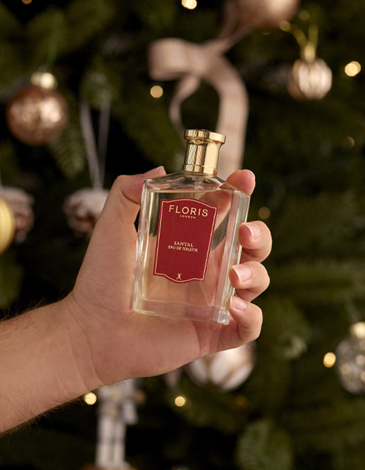 A hand holding a bottle of Floris London Santal fragrance infront of a decorated Christmas Tree. 