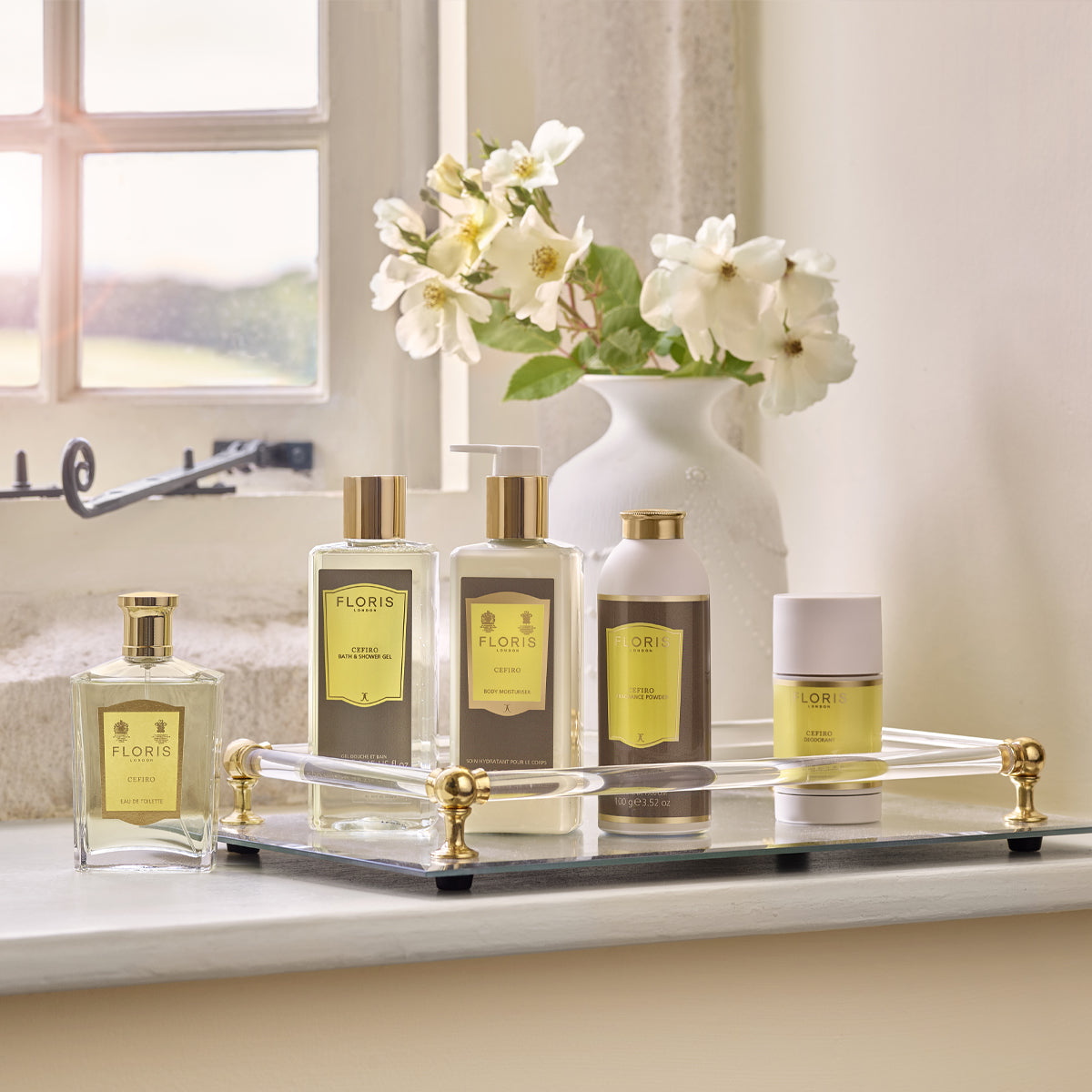 Vegan luxury bath products and Floris London's Cefiro Eau de Toilette on a tray by a window, with flowers in a vase for an elegant touch.