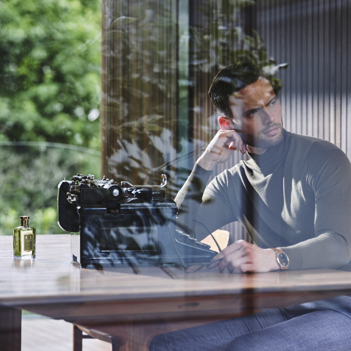 A man in a turtleneck sits by a typewriter and Elite - Eau de Toilette by Floris London, gazing out at greenery as woody citrus notes fill the air.