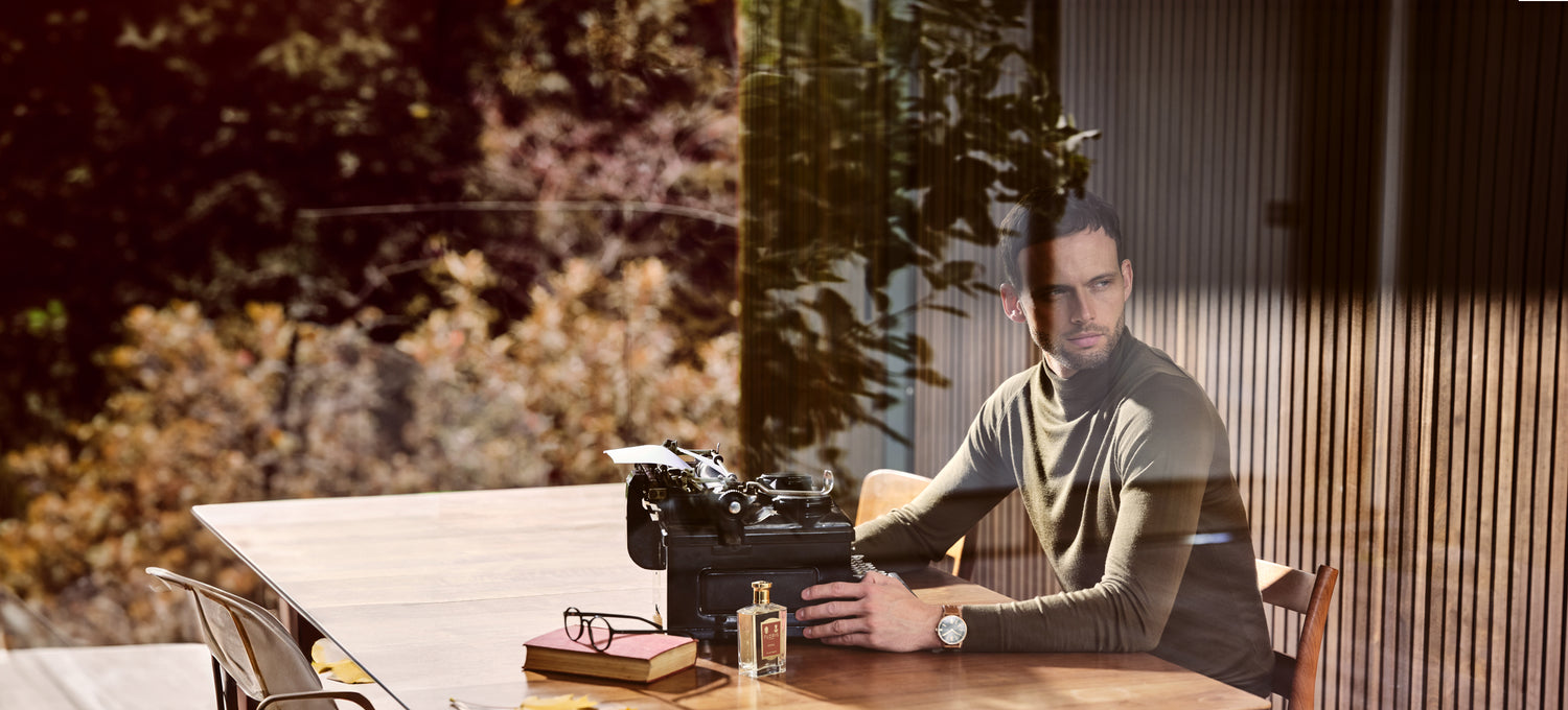 A man sat at a table with a typewriter and a bottle of 'Santal'. 