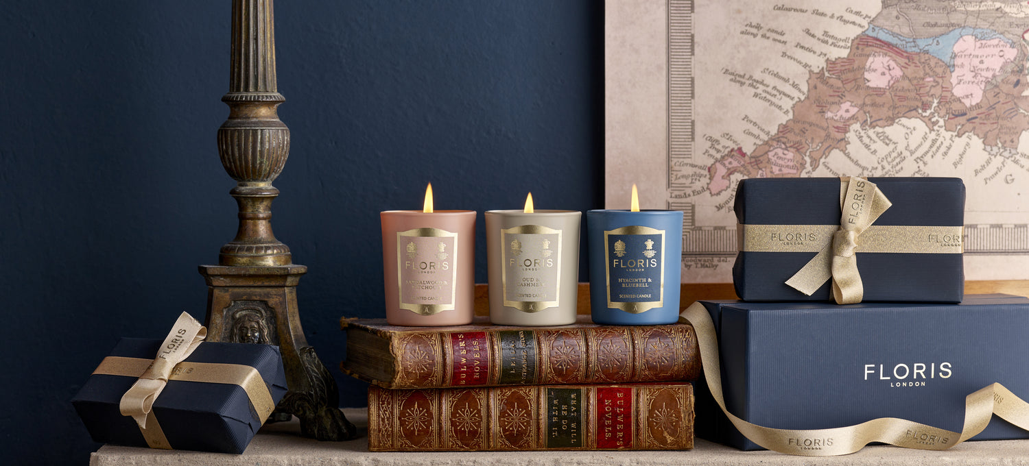 Three beautifully scented candles from the Miniature Candle Collection by Floris London sit on antique books near a decorative map, beside gift-wrapped boxes with ribbons on a mantel.