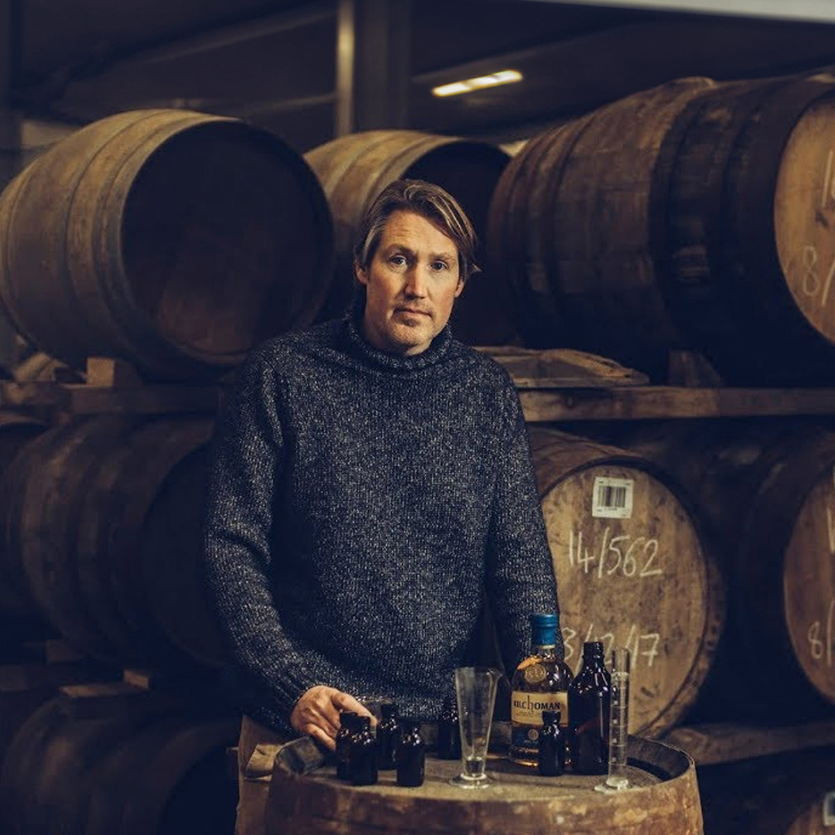 A person stands by whiskey barrels with bottles and a glass on a barrel table, savoring the peaty smoky aroma of Islay by Floris London UK whisky.