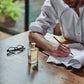 A man writes at a wooden table indoors, with Floris London's JF - Eau de Toilette for men, featuring citrus and woody fragrances, nearby in soft natural light.