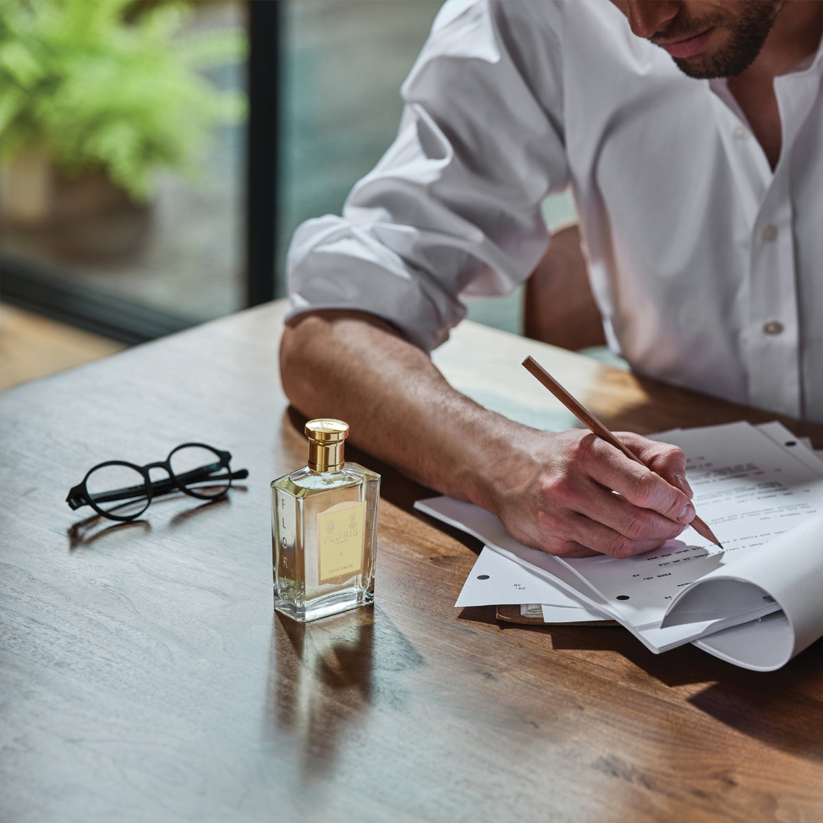 A man writes at a wooden table indoors, with Floris London's JF - Eau de Toilette for men, featuring citrus and woody fragrances, nearby in soft natural light.