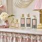 Floris London's Lily Eau de Toilette and body care products are displayed on a ruffled cloth-covered shelf, adorned with flowers and a pink vase.