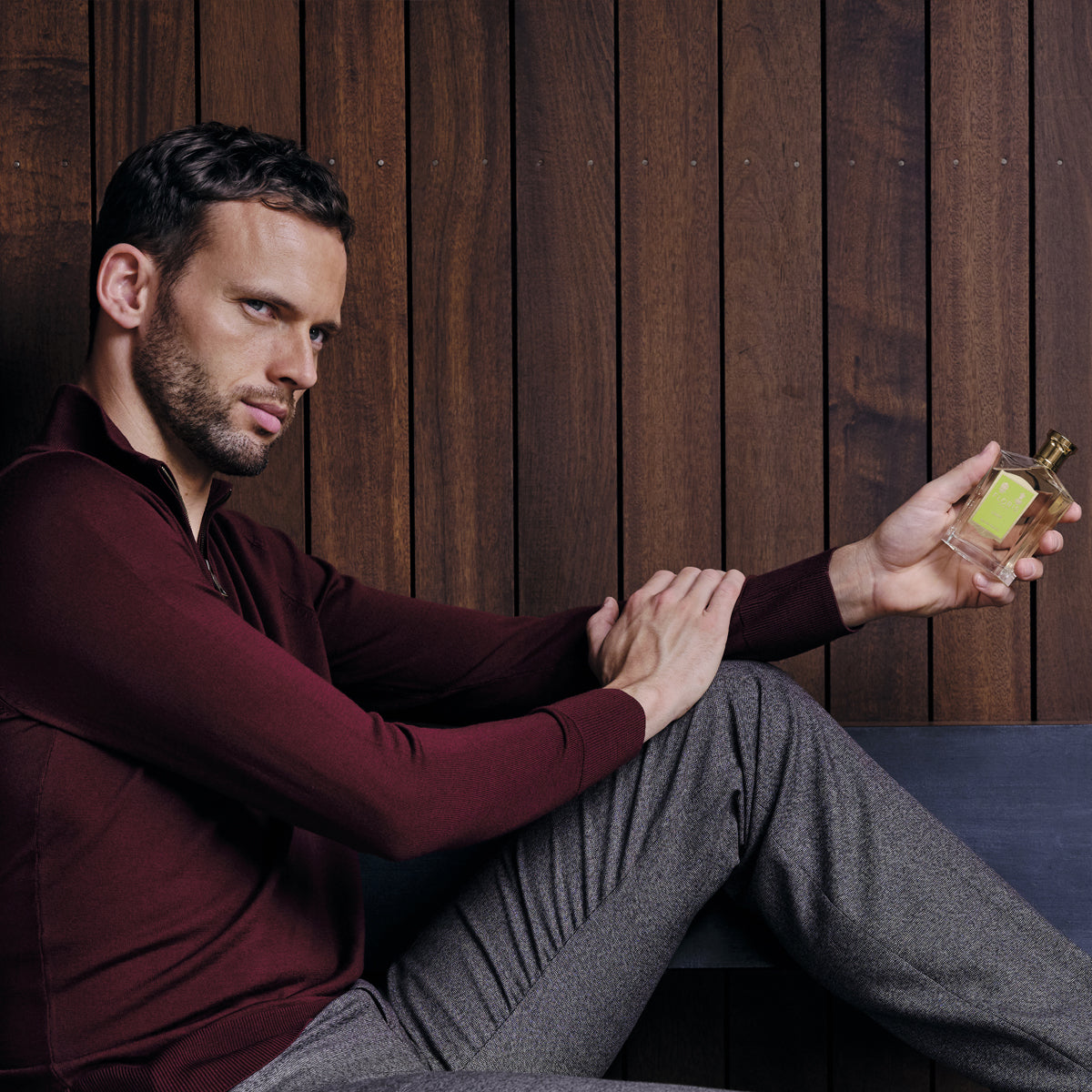 A man in a burgundy sweater and gray pants sits on a bench, showcasing Floris London's Limes Eau de Toilette against a wooden wall.