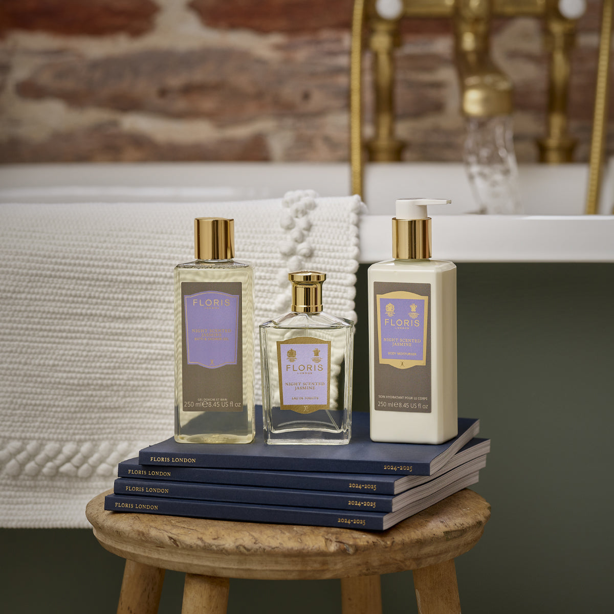 Floris London's Night Scented Jasmine Eau de Toilette sits on books, surrounded by lanterns, with a bathtub and a brick wall backdrop.