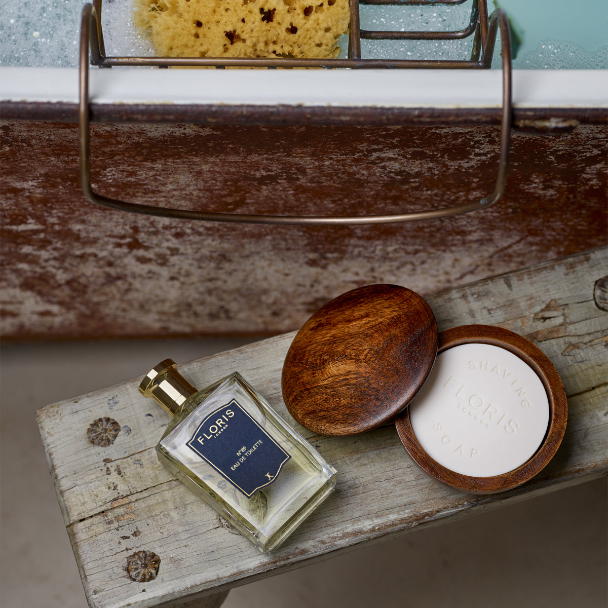 No. 89 - Eau de Toilette by Floris London and classic cologne shaving soap on a rustic stool beside a bubble-filled bath with a sponge.