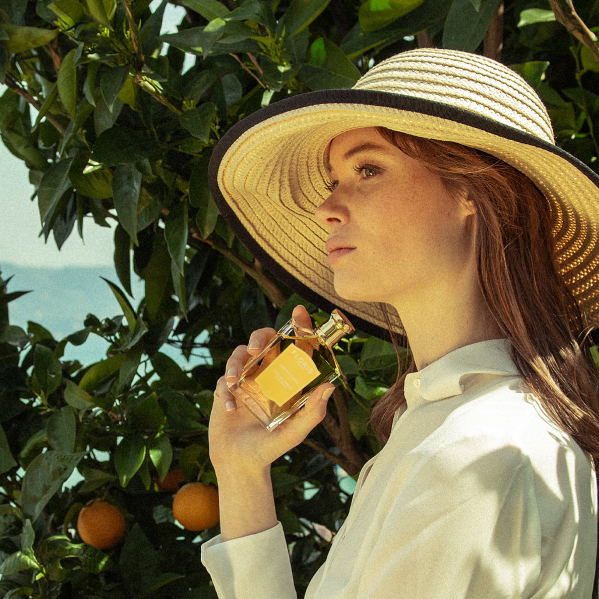 Woman in a sun hat holds a bottle of Floris London UK's Soulle Ámbar jasmine-infused perfume, with citrus trees in the background.