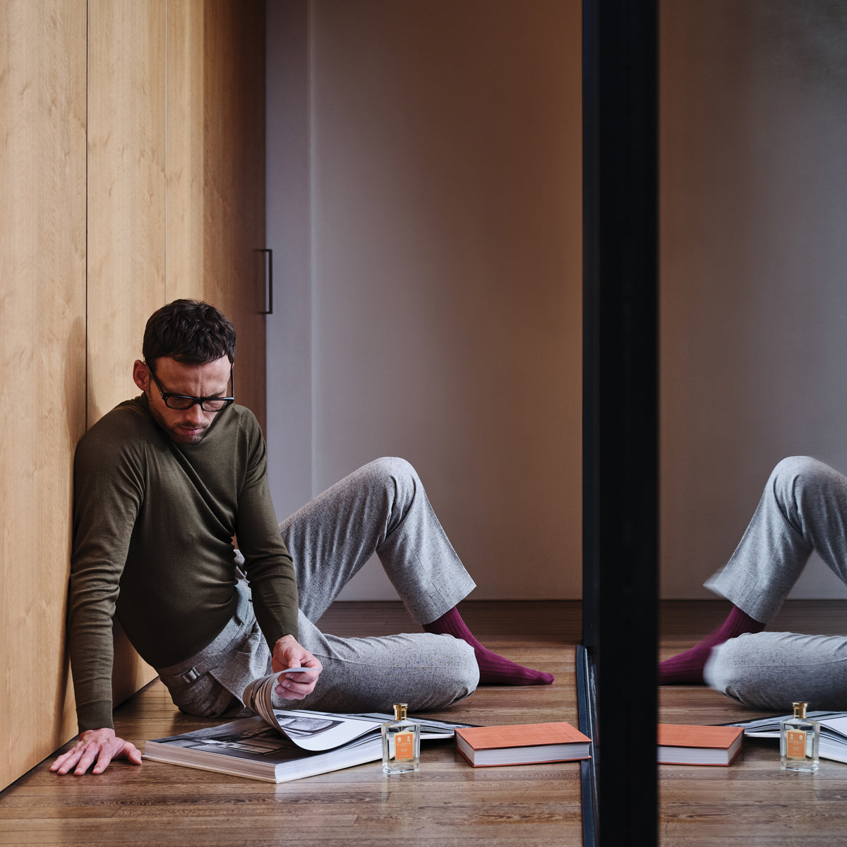 A man reads a magazine on the floor, surrounded by books and a bottle of Floris London's vegan Special No. 127 - Eau de Toilette, reflected in a mirrored wall.