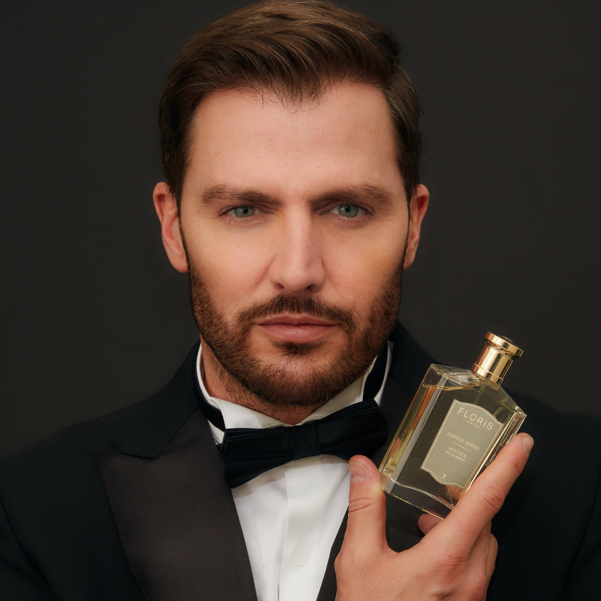 A man in a tuxedo holds a bottle of Floris London UK's Vetiver, with its patchouli fragrance subtly accentuating the dark backdrop.